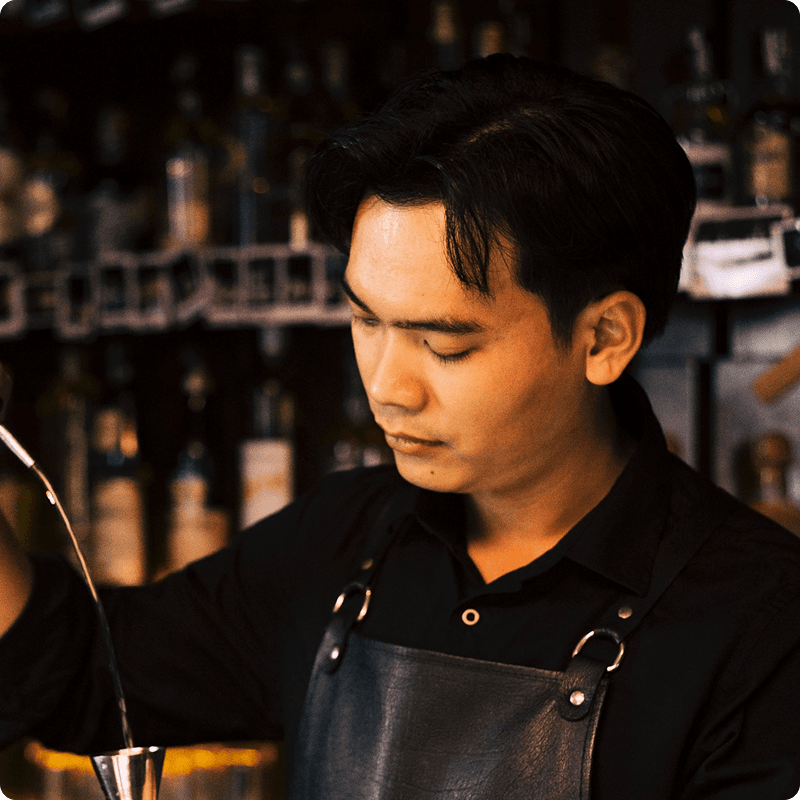Bartender in a dark apron pouring a drink in a dimly lit bar