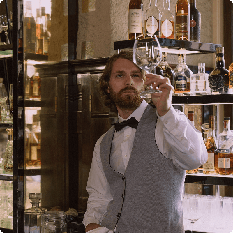 Waiter in a bow tie and waistcoat holding up a glass in front of a bottle shelf