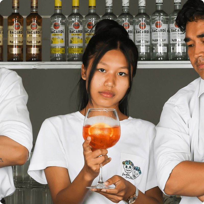 Bar worker in a white T-shirt holding an orange spritz in front of a bottle shelf