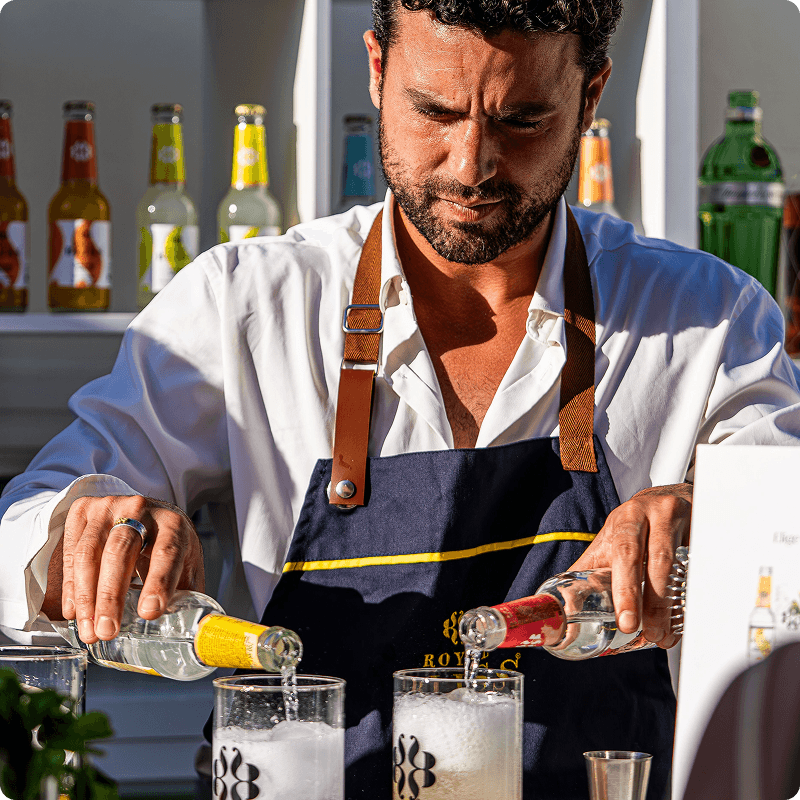 Bartender in a white shirt and apron pouring from two bottles into glasses